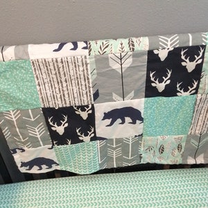 May include: A patchwork baby quilt with a woodland theme. The quilt features a variety of fabrics in shades of gray, blue, and green. The fabrics feature patterns of arrows, trees, deer, and bears.