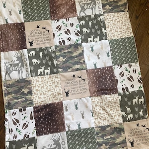 May include: A patchwork baby quilt with a woodland theme. The quilt features a variety of fabrics, including camouflage, brown and white deer print, and a white fabric with brown deer footprints. The quilt also features the text "Ducks, Trucks & Eight Point Bucks That's What Little Boys Are Made Of" and "You Are So Deerly Loved".