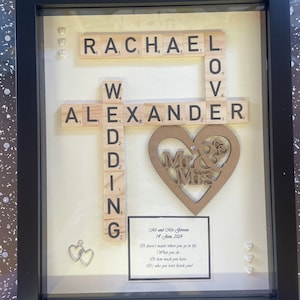 May include: A framed wedding gift with the names "RACHAEL" and "ALEXANDER" spelt out using Scrabble tiles. A wooden heart with "Mr & Mrs" is centered. The frame also includes a printed message and small heart decorations.