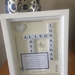 Puede incluir: Un regalo enmarcado para el 25 aniversario de bodas. El marco blanco contiene un fondo color crema con los nombres "Alison" y "Jonathan" escritos con fichas de Scrabble. Los elementos decorativos incluyen un corazón, corazones entrelazados y un poema.