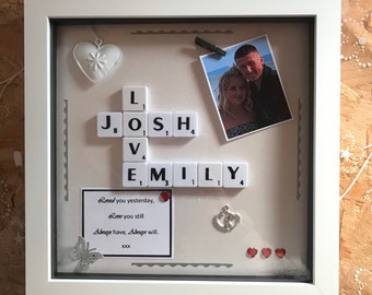 May include: A white framed shadow box with the names "JOSH" and "EMILY" spelled out with Scrabble tiles. The box includes a photo, a heart-shaped ornament, and a small card with text. Decorative elements include a butterfly and hearts.