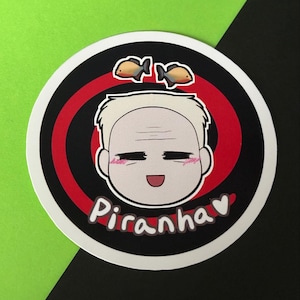 Könnte beinhalten: Runder Aufkleber mit einer Cartoon-Illustration einer Person mit lächelndem Gesicht und dem Wort "Piranha" in weißer Schrift. Zwei kleine Fische befinden sich über dem Kopf. Der Hintergrund hat rote und schwarze konzentrische Kreise.