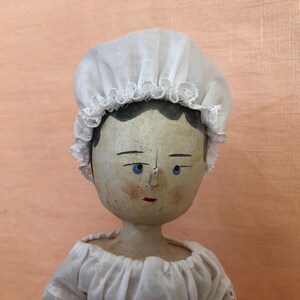 May include: A porcelain doll with a painted face and blue eyes. The doll is wearing a white lace bonnet and a white dress.