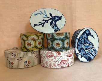 May include: Five oval-shaped boxes with different patterns. The top box has a blue and white deer design. The next box down has a yellow and orange floral pattern. The third box has a blue and white floral pattern. The fourth box has a red and white floral pattern. The bottom box has a white and blue striped pattern with small red and blue flowers.