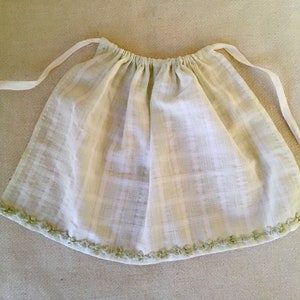 May include: A light green and white plaid apron with a gathered waist and white ties. The bottom edge is trimmed with small green ribbon flowers. The apron is made of a woven fabric.