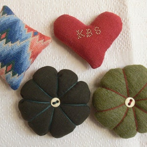 May include: A collection of four fabric pincushions: one diamond-shaped with a blue, pink, and green geometric pattern, one red heart-shaped with the letters KBS embroidered in white, one dark brown flower-shaped, and one olive green flower-shaped.