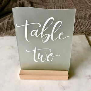 K&ouml;nnte beinhalten: Ein Tischnummernschild aus mattiertem Glas mit einem Holzsockel. Das Schild zeigt "Tisch Zwei" in wei&szlig;er Kalligraphie.