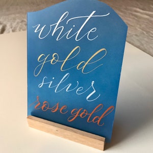 K&ouml;nnte beinhalten: Ein blaues Schild mit den Worten "white gold silver rose gold" in verschiedenen Farben geschrieben. Das Schild steht auf einem Holzst&auml;nder.