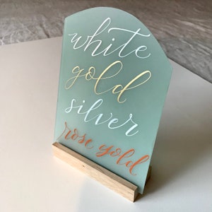K&ouml;nnte beinhalten: Ein mattes Glasschild mit einem Holzst&auml;nder. Auf dem Schild stehen die Worte "Wei&szlig;gold", "Silber" und "Ros&eacute;gold" in verschiedenen Farben.