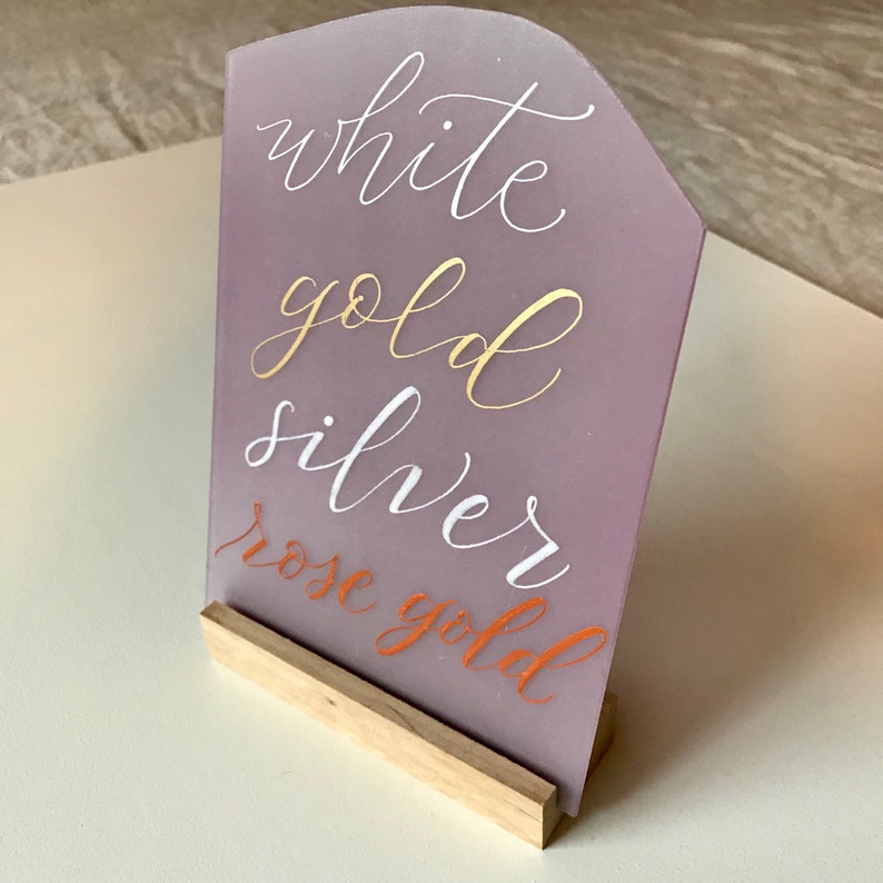 K&ouml;nnte beinhalten: Ein lilafarbenes Acrylschild mit einem Holzst&auml;nder. Das Schild listet die folgenden Metalloberfl&auml;chen auf: Wei&szlig;gold, Silber und Ros&eacute;gold.