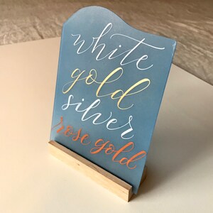 K&ouml;nnte beinhalten: Ein blaues Acrylschild mit einem Holzst&auml;nder. Das Schild tr&auml;gt die Aufschrift "white gold silver rose gold" in goldener, wei&szlig;er und orangefarbener Farbe.