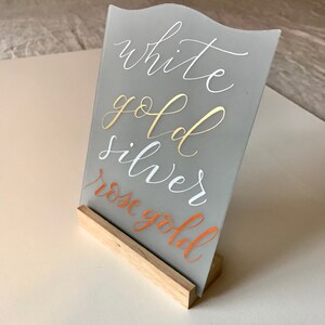 K&ouml;nnte beinhalten: Ein graues Acrylschild mit einem Holzst&auml;nder. Auf dem Schild stehen die Worte "white gold silver rose gold" in verschiedenen Farben von Metallic-Farbe.