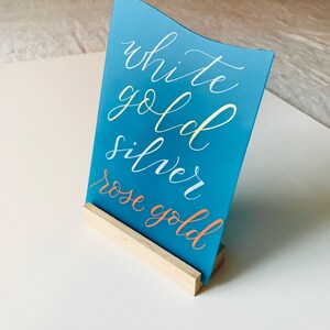 K&ouml;nnte beinhalten: Ein t&uuml;rkisblaues Schild mit wei&szlig;er Kalligraphie, auf dem "white gold silver rose gold" steht, auf einem Holzst&auml;nder.