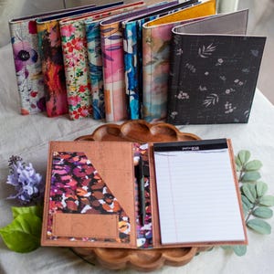 May include: A collection of upright rectangular notebook covers featuring various floral and abstract patterns in vibrant colors. One open cover displays a tan interior with a patterned pocket, a black pen, and a white lined Pen+Gear notepad.