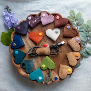 May include: A selection of heart-shaped cork cable tidies in diverse hues, each featuring a coloured button fastening. A black USB cable is neatly arranged within one, presented on a wooden tray with floral decorations.