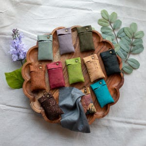 May include: A collection of small rectangular pouches made from cork material in various colors including green, purple, olive, red, lime, tan, black, turquoise, and brown with metallic speckles, each featuring a brass snap button and a metal ring.