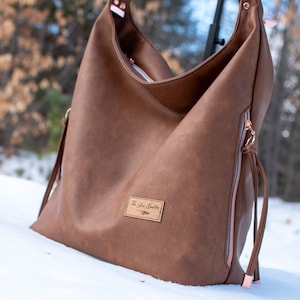 May include: A brown hobo bag with a soft, textured finish. The bag has a rose gold zip and a small leather patch with text. It features a shoulder strap and is set against a snowy backdrop.
