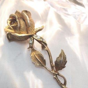 May include: A gold-toned rose brooch with a textured finish. The brooch features a detailed rose bloom, leaves, and a stem. Small, clear rhinestones accent the stem. The brooch is set against a white, textured background.