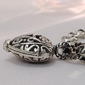 May include: Close-up of a silver-toned, ornate bead with intricate scrollwork and a textured surface. The bead is part of a chain, with other decorative elements visible. The design suggests a jewelry component.