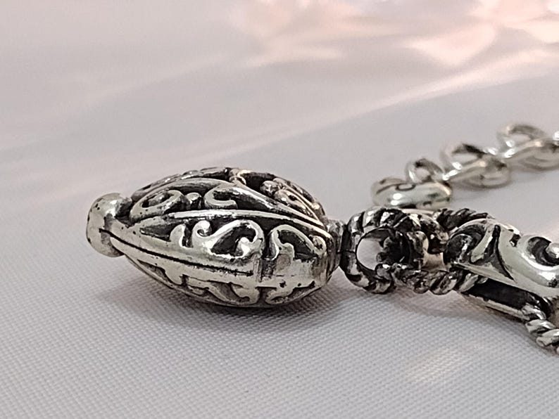 May include: Close-up of a silver-toned, ornate bead with intricate scrollwork and a textured surface. The bead is part of a chain, with other decorative elements visible. The design suggests a jewelry component.