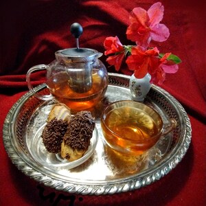 May include: A silver tray with a glass teapot filled with tea, a cup of tea, and two chocolate-covered cookies. The tray is decorated with a floral design and is sitting on a red tablecloth. There are pink flowers in a small white vase on the tray.