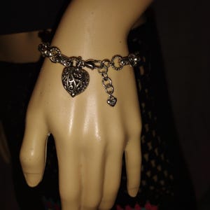 May include: Silver-toned bracelet featuring a heart-shaped charm with an intricate openwork design. The bracelet has a chain-link style with a lobster clasp closure and a small heart charm dangling near the clasp. The bracelet is displayed on a mannequin hand.