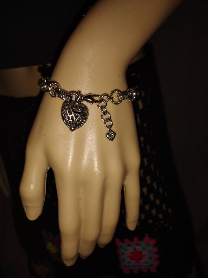 May include: Silver-toned bracelet featuring a heart-shaped charm with an intricate openwork design. The bracelet has a chain-link style with a lobster clasp closure and a small heart charm dangling near the clasp. The bracelet is displayed on a mannequin hand.