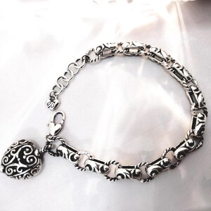May include: Silver-toned bracelet with an ornate, scrolling design. The bracelet features a heart-shaped charm with a similar filigree pattern. The adjustable chain and clasp are also silver, creating a cohesive and elegant piece of jewelry.