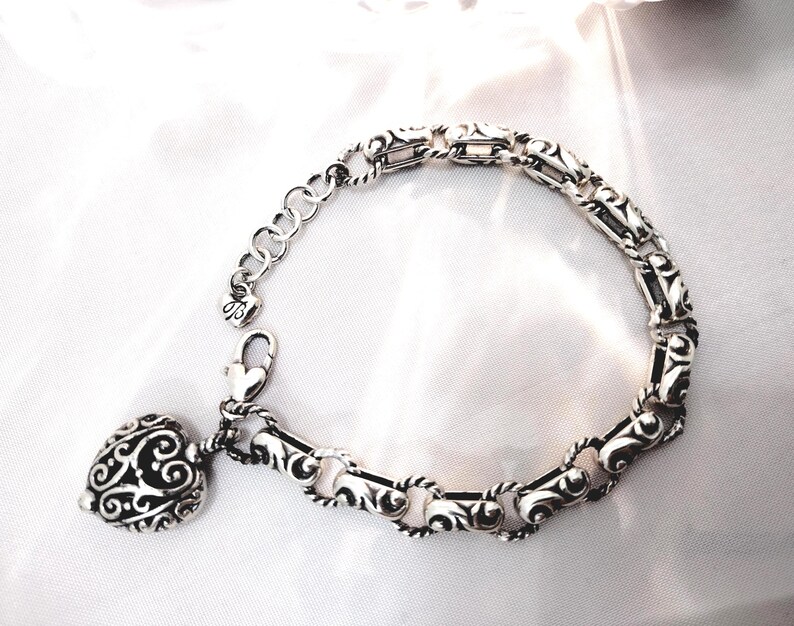 May include: Silver-toned bracelet with an ornate, scrolling design. The bracelet features a heart-shaped charm with a similar filigree pattern. The adjustable chain and clasp are also silver, creating a cohesive and elegant piece of jewelry.