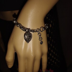 May include: Silver-toned bracelet with a heart-shaped charm. The charm has an open filigree design. The bracelet is on a mannequin hand. The bracelet has a lobster claw clasp and an adjustable chain.