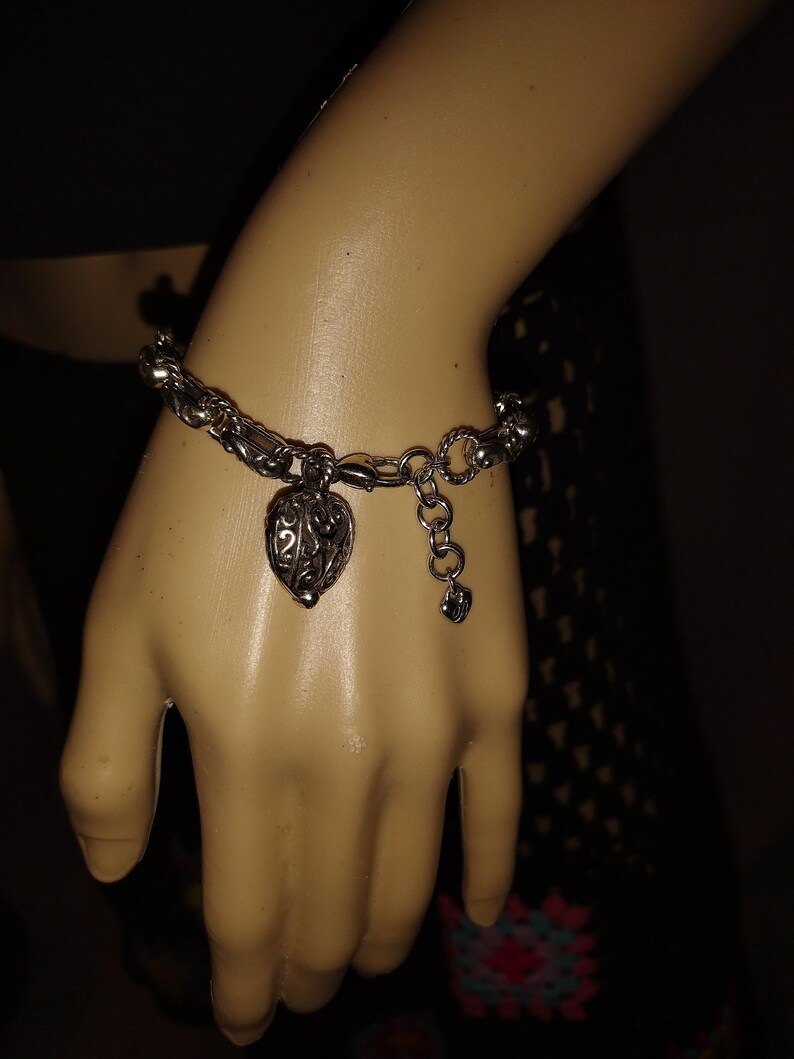 May include: Silver-toned bracelet with a heart-shaped charm. The charm has an open filigree design. The bracelet is on a mannequin hand. The bracelet has a lobster claw clasp and an adjustable chain.