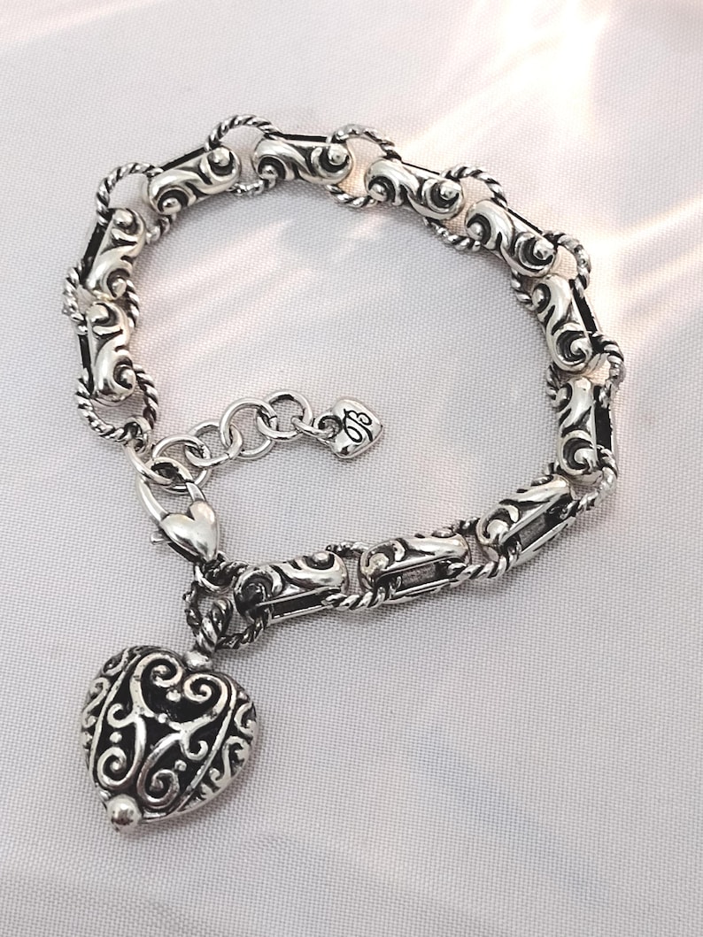 May include: Silver bracelet with a heart-shaped charm. The bracelet features an ornate design with a heart pendant. The charm is detailed with a filigree pattern. The bracelet is made of silver and has a lobster clasp closure.