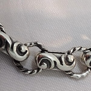 May include: Close-up of a silver chain necklace with ornate, scroll-like links. The links are connected by twisted rope-like sections. The necklace is displayed on a light-colored, textured surface.