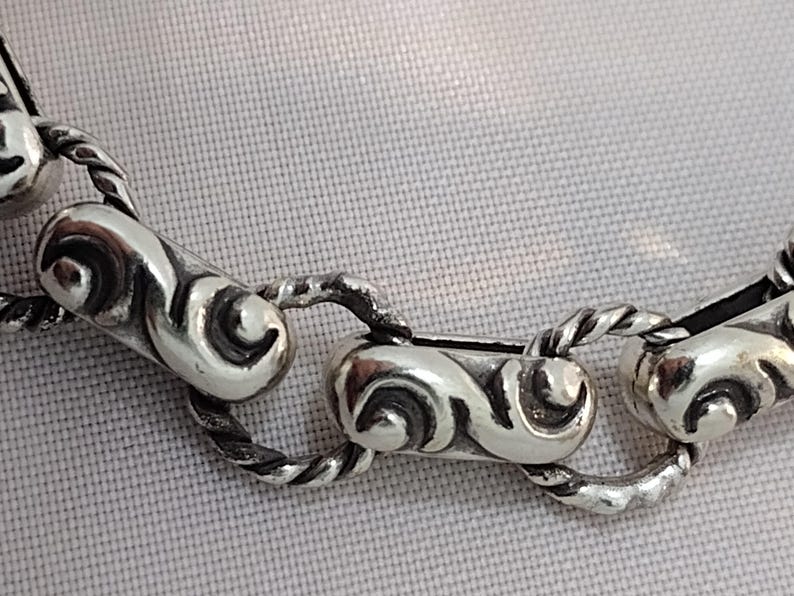 May include: Close-up of a silver chain necklace with ornate, scroll-like links. The links are connected by twisted rope-like sections. The necklace is displayed on a light-colored, textured surface.