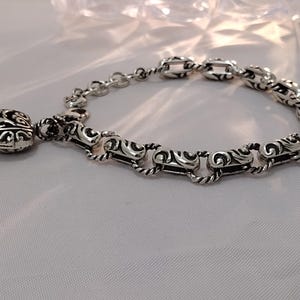 May include: A silver bracelet with an ornate design. The bracelet features a series of interconnected links with a detailed, swirling pattern. A decorative charm with a similar design hangs from the bracelet, adding a touch of elegance.
