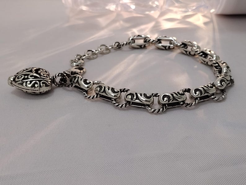 May include: A silver bracelet with an ornate design. The bracelet features a series of interconnected links with a detailed, swirling pattern. A decorative charm with a similar design hangs from the bracelet, adding a touch of elegance.
