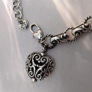 May include: Silver-toned heart pendant necklace with an ornate, openwork design. The heart charm hangs from a chain with a lobster clasp. The necklace features a small heart charm near the clasp.