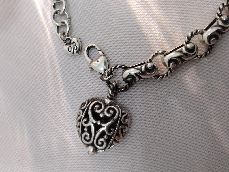 May include: Silver-toned heart pendant necklace with an ornate, openwork design. The heart charm hangs from a chain with a lobster clasp. The necklace features a small heart charm near the clasp.
