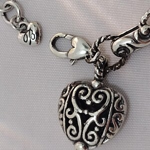 May include: Close-up of a silver-toned heart-shaped pendant with intricate scrollwork. The pendant is attached to a chain with a lobster clasp. A small heart charm with initials is also visible.