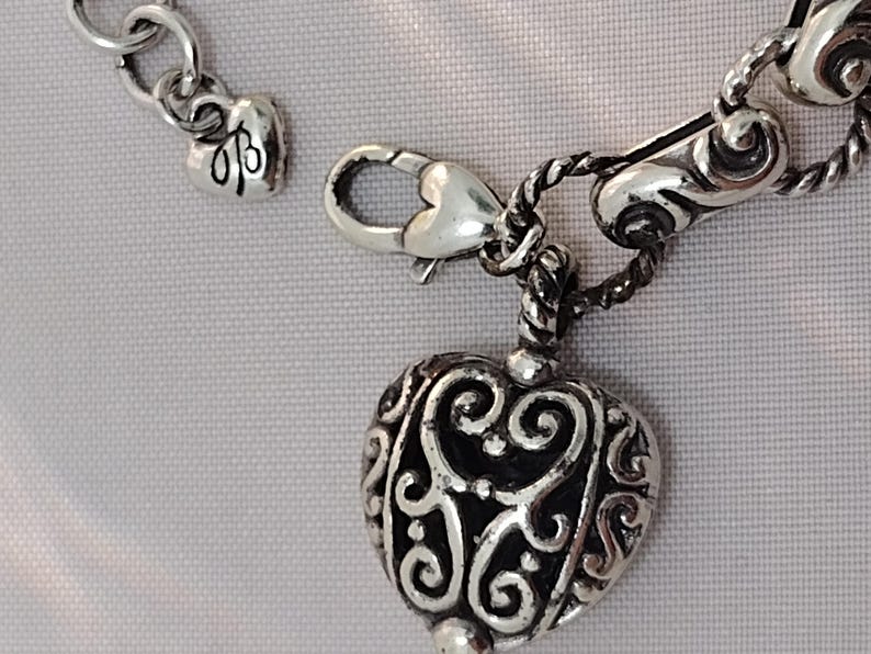 May include: Close-up of a silver-toned heart-shaped pendant with intricate scrollwork. The pendant is attached to a chain with a lobster clasp. A small heart charm with initials is also visible.