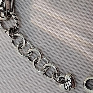 May include: Close-up of a silver-toned bracelet with a decorative clasp. The bracelet features a series of circular links and a heart-shaped charm with an engraved design. The background is a light gray fabric.