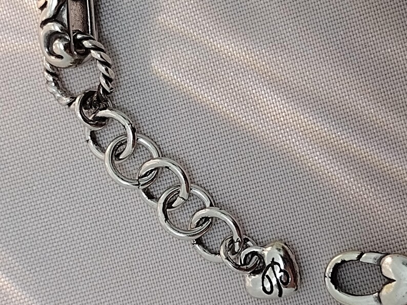 May include: Close-up of a silver-toned bracelet with a decorative clasp. The bracelet features a series of circular links and a heart-shaped charm with an engraved design. The background is a light gray fabric.