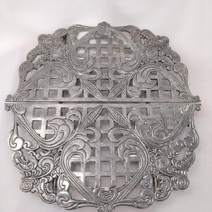 May include: A silver-colored metal trivet featuring a symmetrical, intricate openwork design. The piece is composed of four square grid sections surrounded by ornate, swirling scrollwork patterns, creating a circular overall shape.