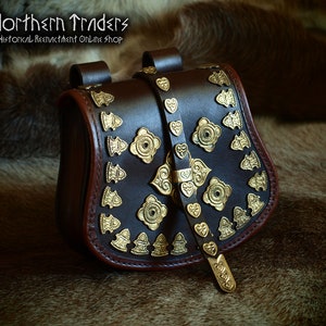 Op de afbeelding: Een bruine leren tas met een gouden metalen gesp en decoratieve accenten. De tas heeft een band om hem over de schouder te dragen. "Northern Traders Historical Reenactment Online Shop" staat bovenaan de tas gedrukt.