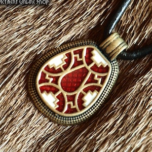 May include: A teardrop-shaped pendant with a brass-toned metal frame featuring a textured border. The center contains a red enamel inlay with white geometric patterns. It hangs from a black cord. The text Northern Traders Historical Reenactment Online Shop is visible in the top left corner.