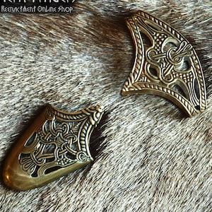 May include: Two brass-colored metal fittings featuring intricate, openwork knotwork patterns. Each piece has a curved, triangular shape with a smooth, solid border and a detailed, recessed center design. The metal has a polished, antique finish.