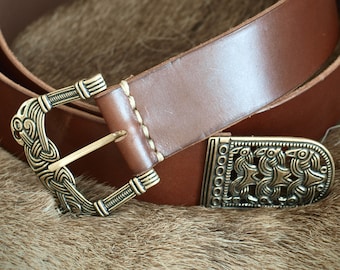 May include: A brown leather belt with a decorative brass buckle featuring a Viking knot design. The buckle is ornate and has a detailed pattern. The belt is folded in half and the buckle is closed.