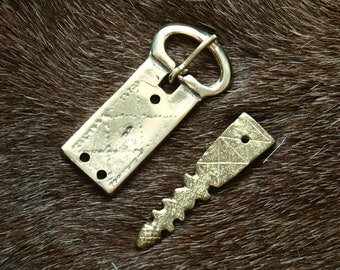 May include: A gold-colored metal belt buckle with a rectangular plate and a separate, ornate, pointed tongue. The buckle is on a brown fur background.