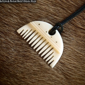 Puede incluir: Un peine de hueso blanco con un diseño decorativo y un cordón de cuero negro. El peine tiene forma de semicírculo y tiene un agujero en la parte superior para el cordón. El peine está sobre un fondo de piel marrón.