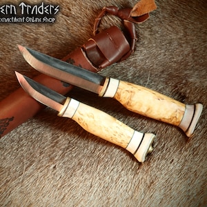 Könnte beinhalten: Zwei Messer mit schwarzen Griffen, Holzgriffen und einer Lederscheide. Die Messer haben eine dunkelbraune Oberfläche und sind aus Metall. Die Scheide hat einen braunen Lederriemen und ein dekoratives Design.